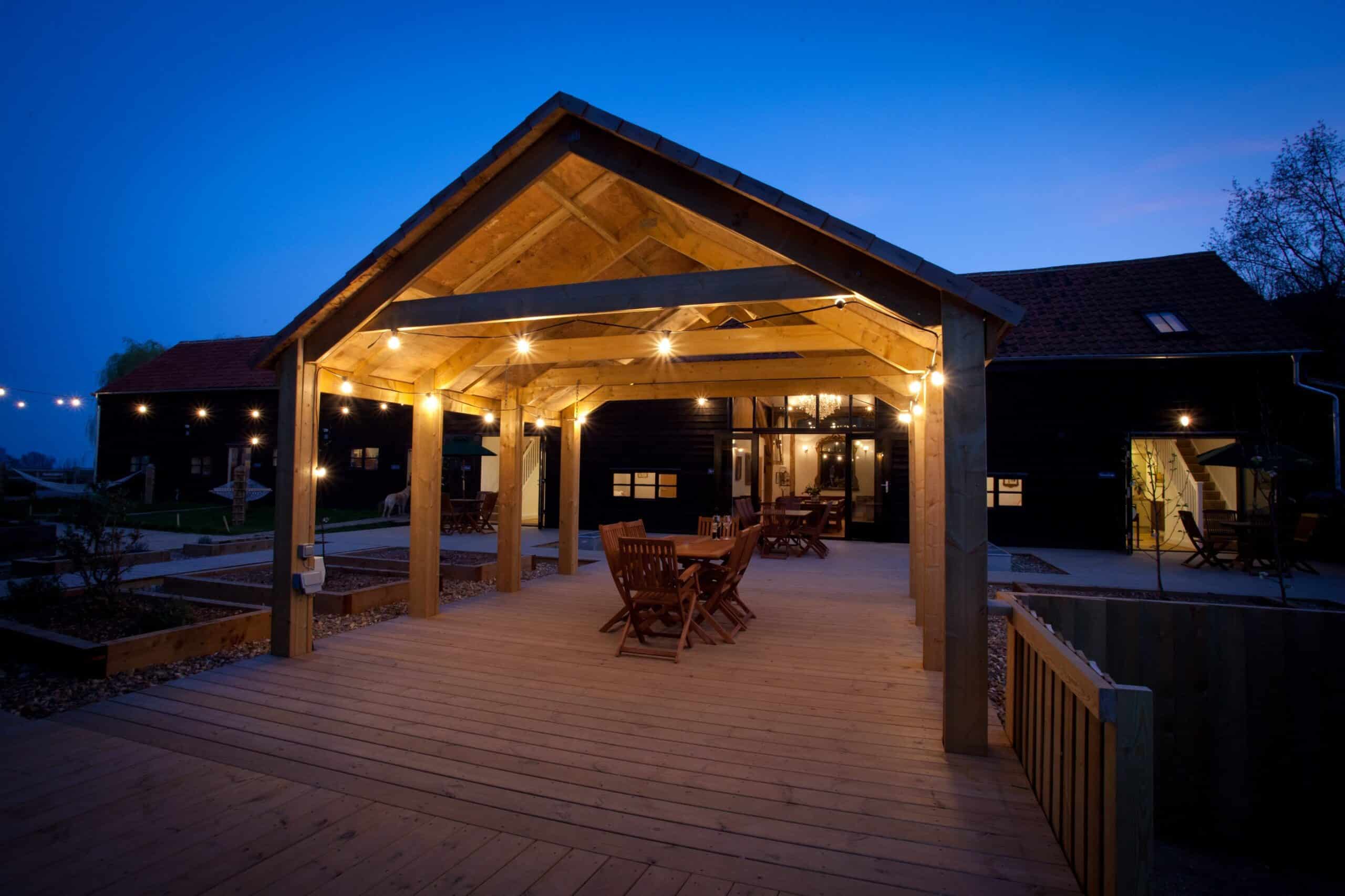 A wooden patio with tables and chairs is lit by fairy lights under a large wooden pergola, attached to a dark accommodation building at dusk. The deep blue sky and tranquil surroundings create an inviting atmosphere. | The Canary Shed