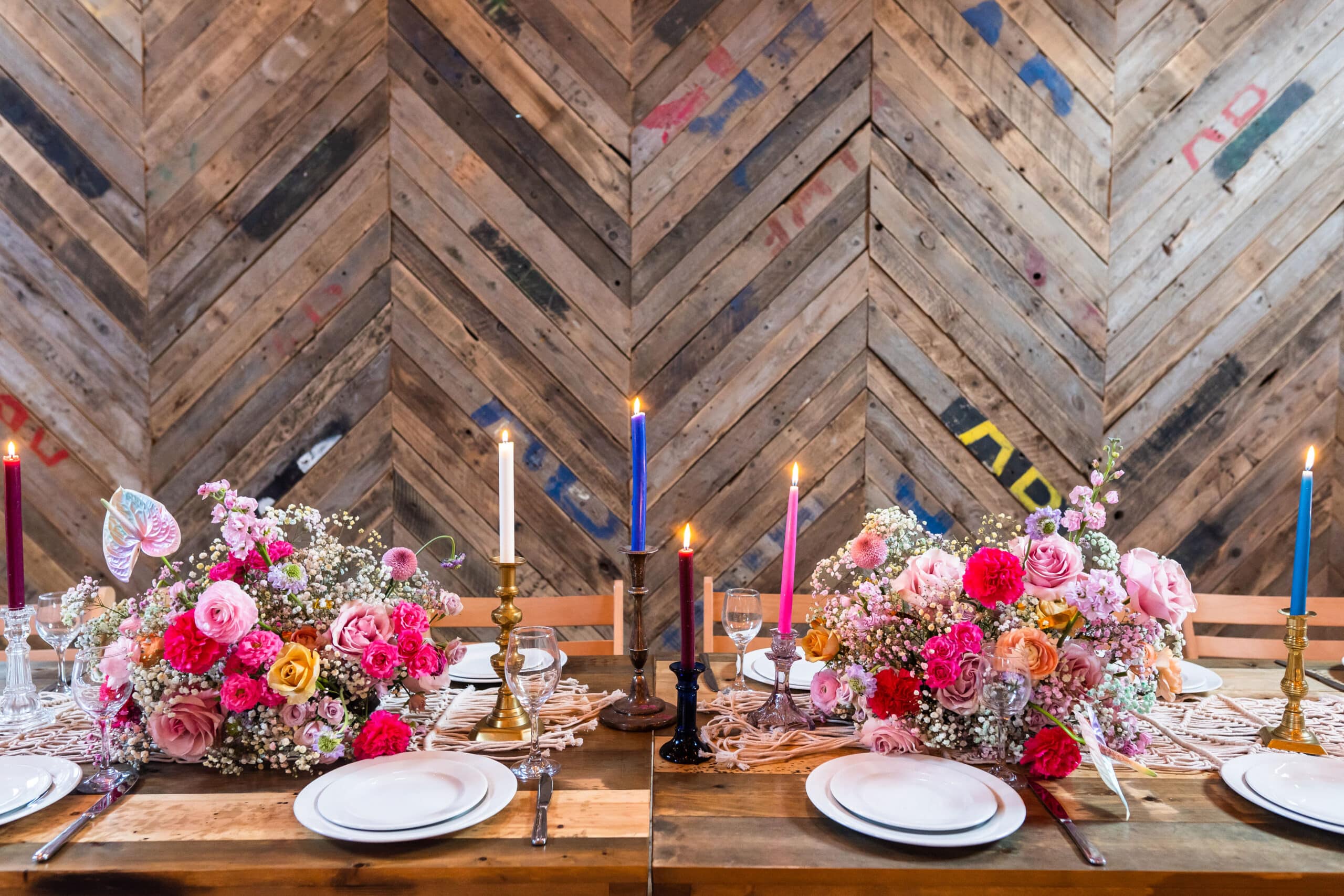 A rustic wooden table is set with white plates, colourful candles, glasses, and vibrant floral centrepieces of pink and red flowers, illustrating how it works to create a warm gathering space against a chevron-patterned wooden wall. | The Canary Shed