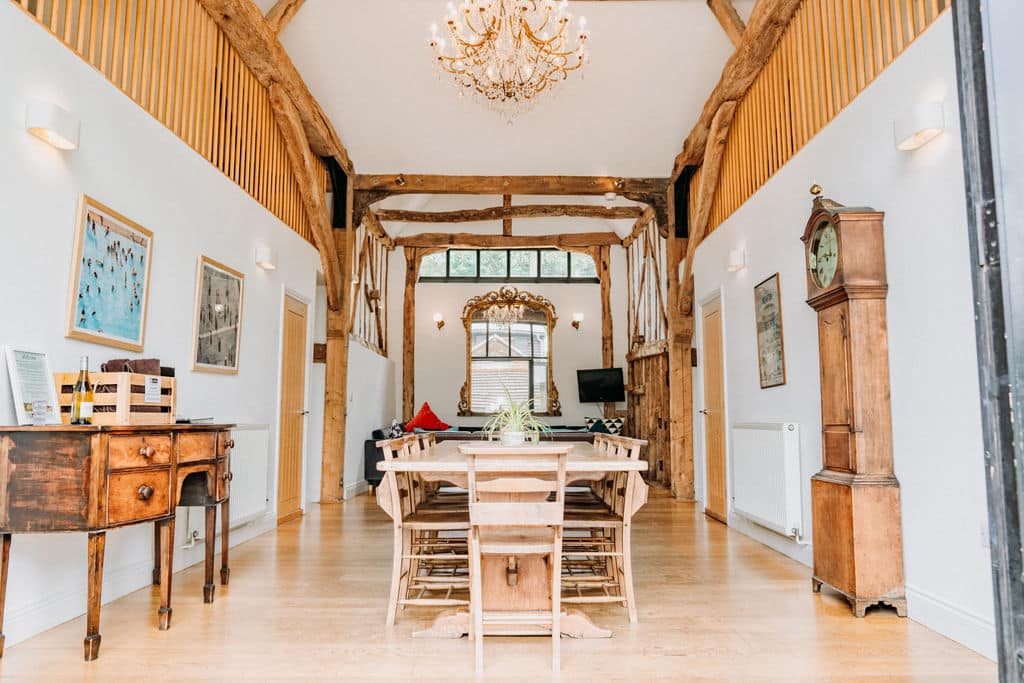 A spacious, rustic dining room with exposed wooden beams, a long wooden table with chairs, chandelier, antique furniture, and grandfather clock. Large windows let in natural light. What's Included: exquisite artwork on white walls. | The Canary Shed