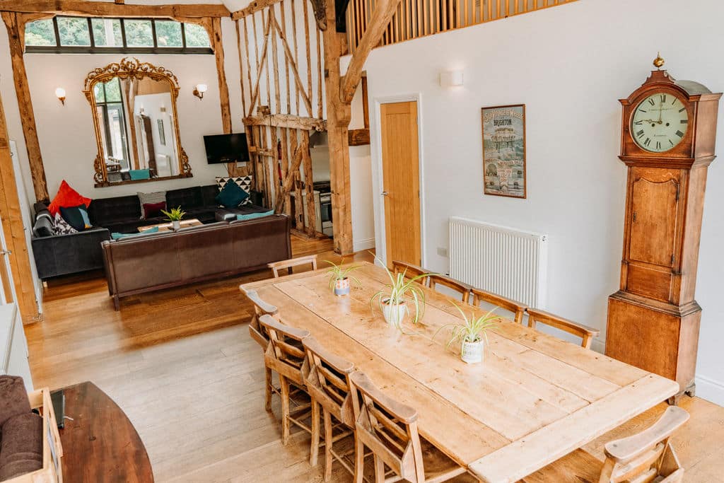 A bright, rustic accommodation featuring a living and dining area with exposed wooden beams, a long wooden dining table adorned with plants, a classic grandfather clock, cosy sofas with cushions, and a large ornate mirror. | The Canary Shed
