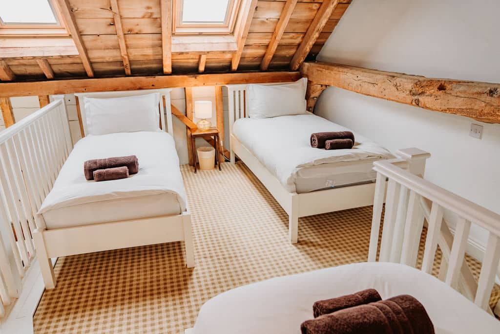 This cosy attic accommodation features two single beds with white bed linen and brown towels, set under a slanted wooden ceiling with rooflights. A small bedside table with a lamp sits between the beds, creating a warm and inviting retreat. | The Canary Shed