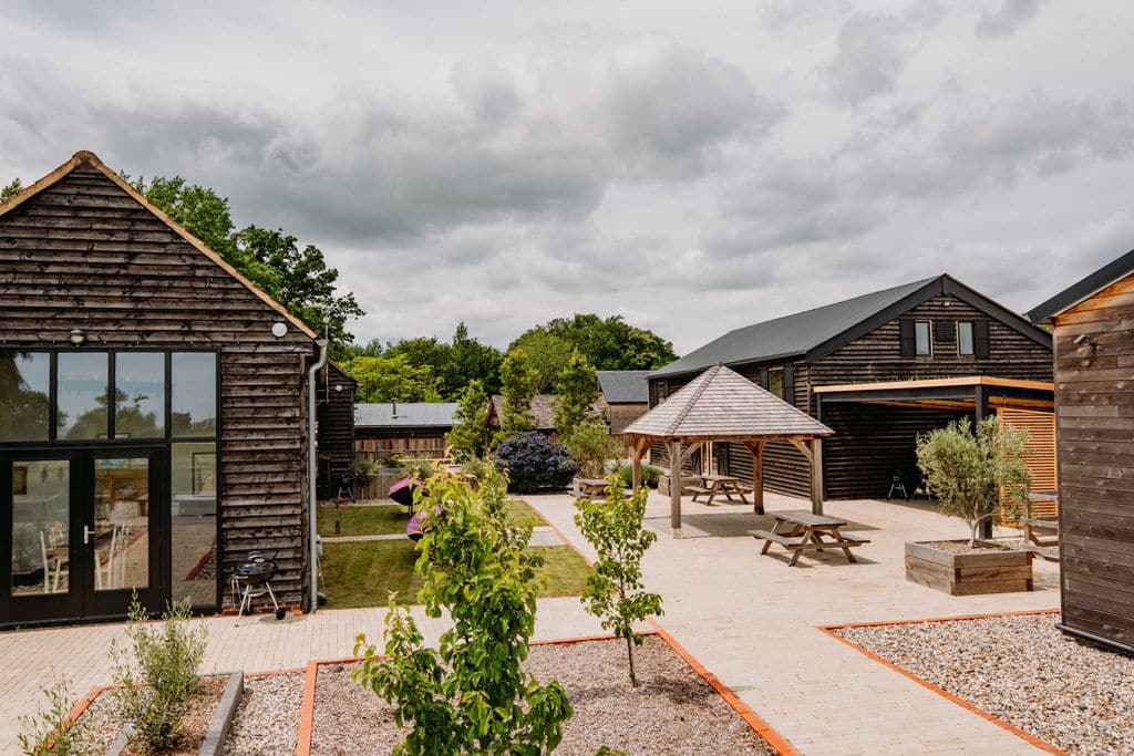 A courtyard with wooden buildings offers unique accommodation; picnic tables rest under a wooden shelter amid young trees and landscaped garden beds beneath a cloudy sky. | The Canary Shed