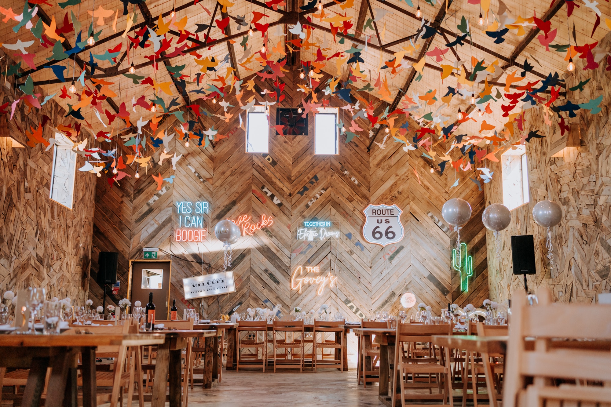 A rustic event hall decorated with colourful hanging paper birds, neon signs, balloons, and long wooden tables set for a celebration—natural light streams in as guests gather to see how it works. | The Canary Shed