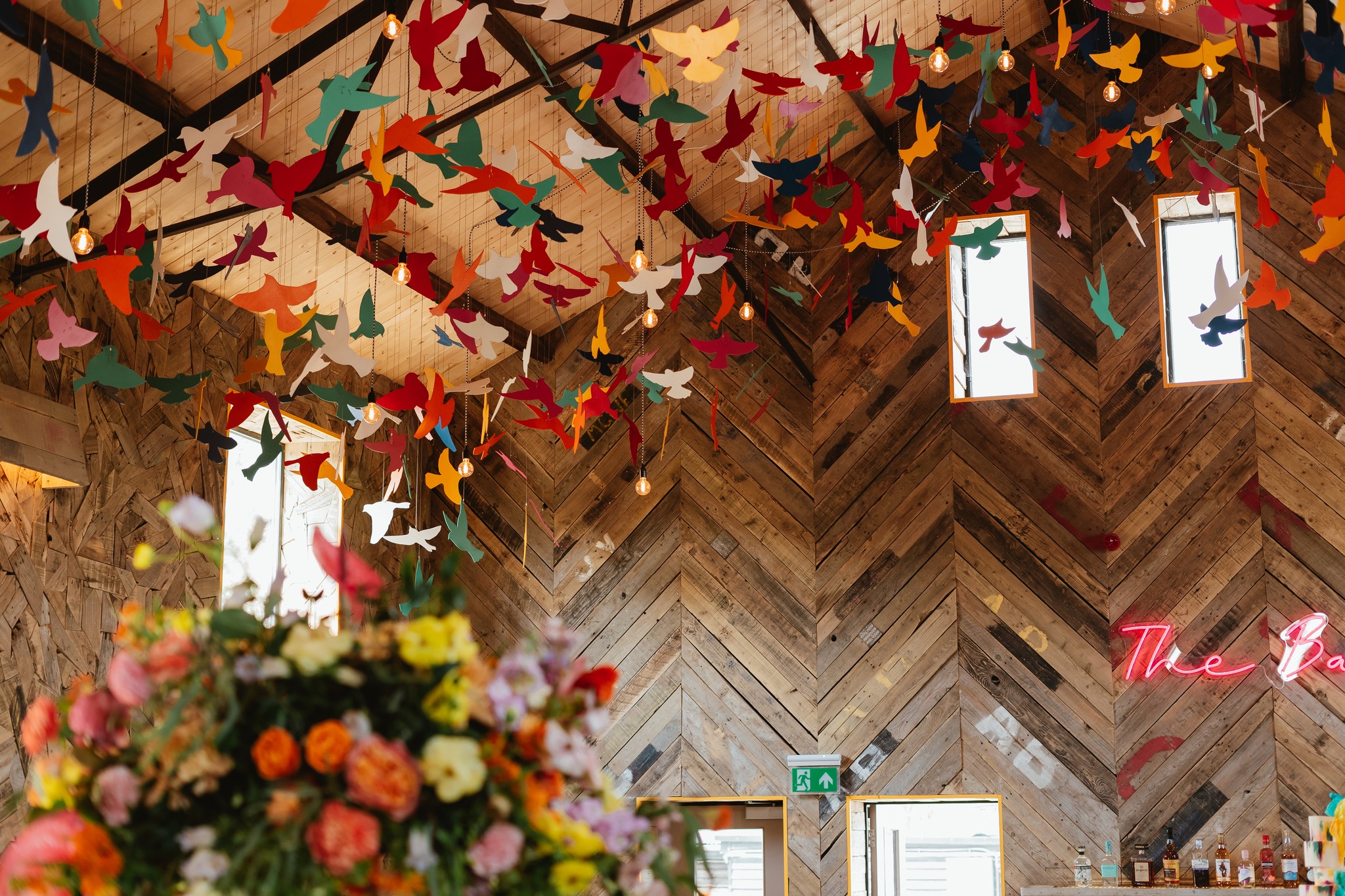 A wooden room with colourful paper birds hanging from the ceiling, a bouquet of flowers in the foreground, and neon signage on the wall. Sunlight streams through tall windows—perfect inspiration for unforgettable wedding planning. | The Canary Shed