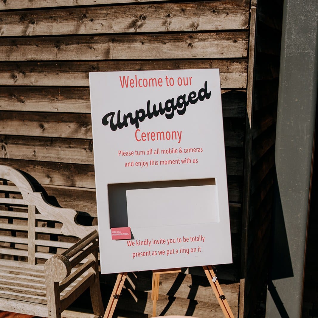 A sign at The Canary Shed reads: Welcome to our Unplugged Ceremony. Please switch off all mobiles & cameras and enjoy this unforgettable wedding moment. Be completely present as we put a ring on it. The sign is on an easel by a wooden wall—one of our top wedding tips!. | The Canary Shed