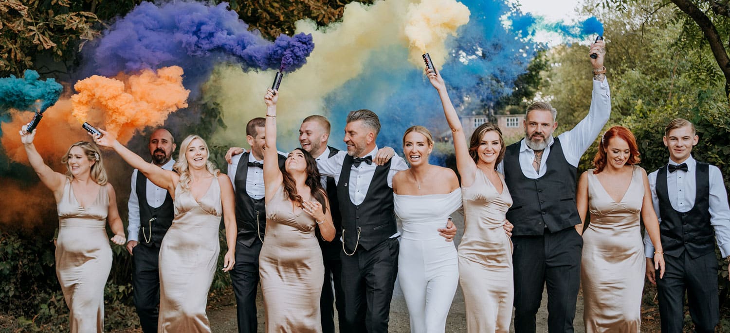 A wedding party of six women in gold dresses and five men in black suits walks outdoors at The Canary Shed, smiling and holding colourful smoke bombs in purple, orange, and yellow. Trees line the path in the background. | The Canary Shed