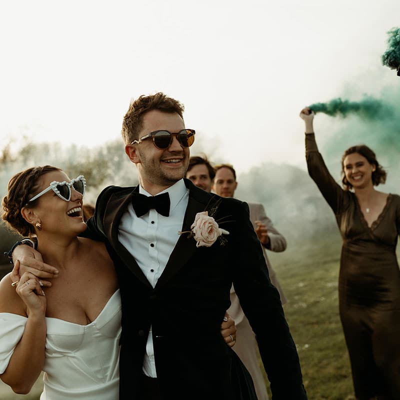 A joyful bride and groom in formal attire smile and embrace outdoors at The Canary Shed, surrounded by friends. One guest waves vibrant smoke flares, adding to the festive atmosphere. The bride wears heart-shaped sunglasses. | The Canary Shed