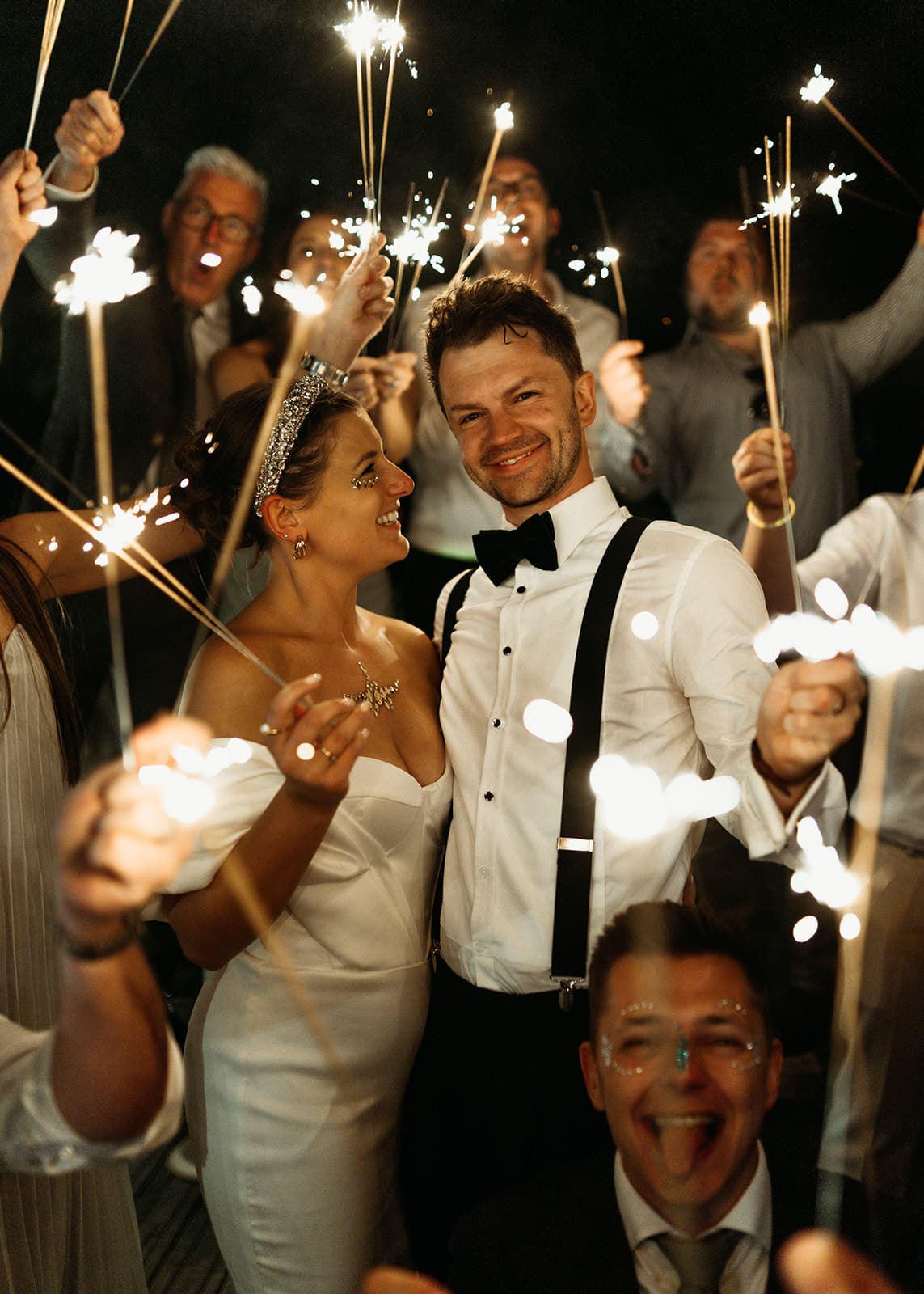 A bride and groom, dressed in wedding attire, smile at each other whilst guests surround them holding sparklers. The scene is joyful and festive, with warm lighting and happy faces celebrating at night. | The Canary Shed