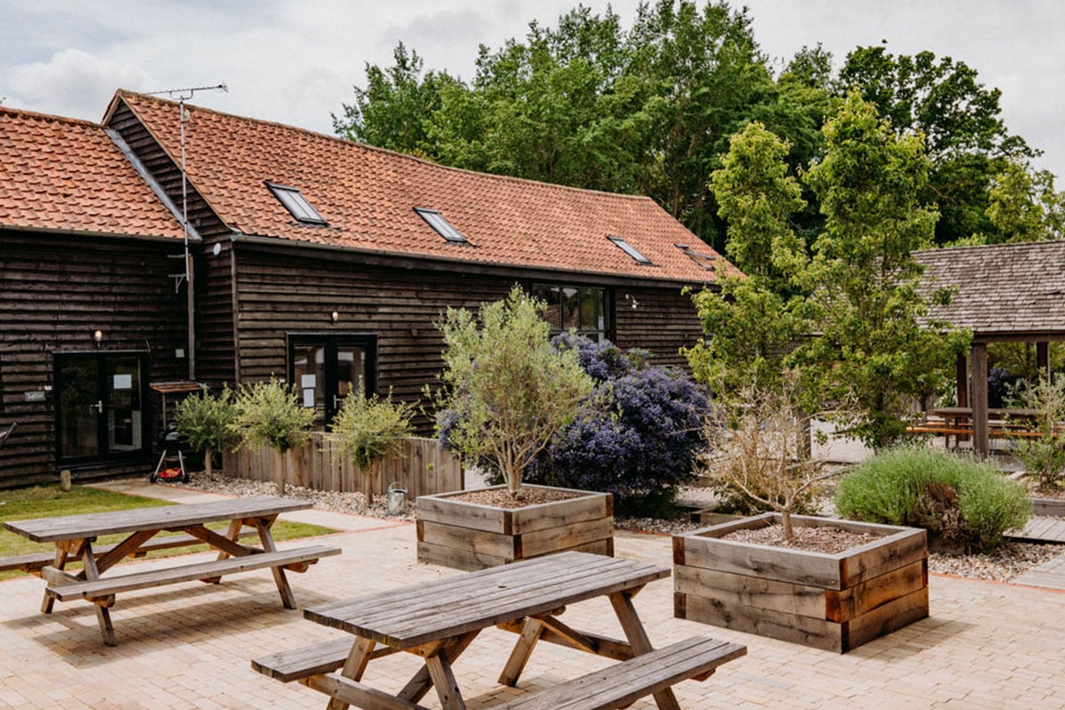 Wooden picnic tables on a brick patio with large planter boxes, trees, and shrubs create an inviting space perfect for spring weddings. A rustic, dark wooden building with a red tiled roof and skylights stands in the background, surrounded by lush greenery. | The Canary Shed