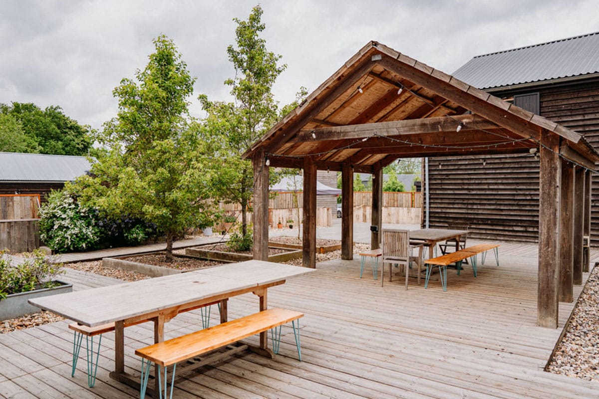 Outdoor wooden decking with picnic tables and benches under a rustic wooden pavilion, surrounded by trees and modern wooden buildings, on a cloudy day—an inviting setting perfect for autumn weddings. | The Canary Shed