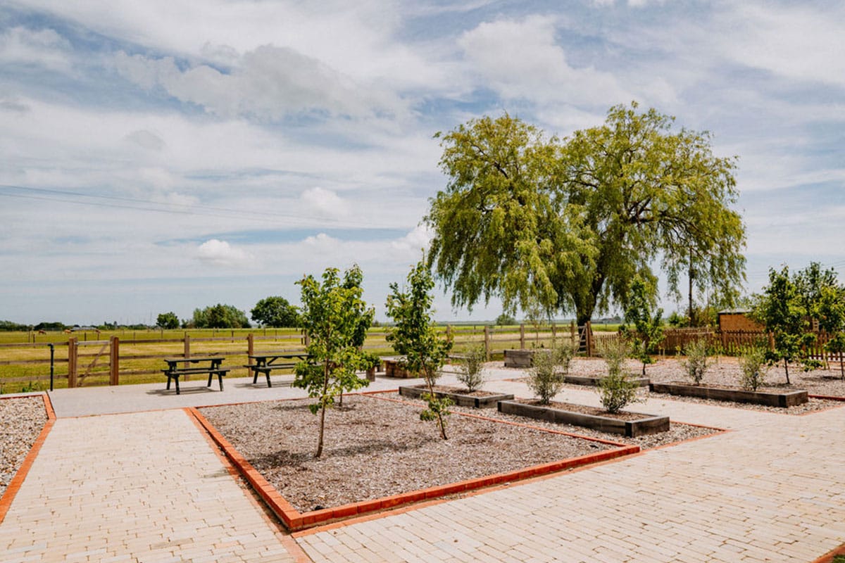 A landscaped garden with raised beds, young trees, a mature tree in the background, two picnic tables, and a wooden fence overlooking a grassy field under a partly cloudy sky—an idyllic setting perfect for summer weddings. | The Canary Shed